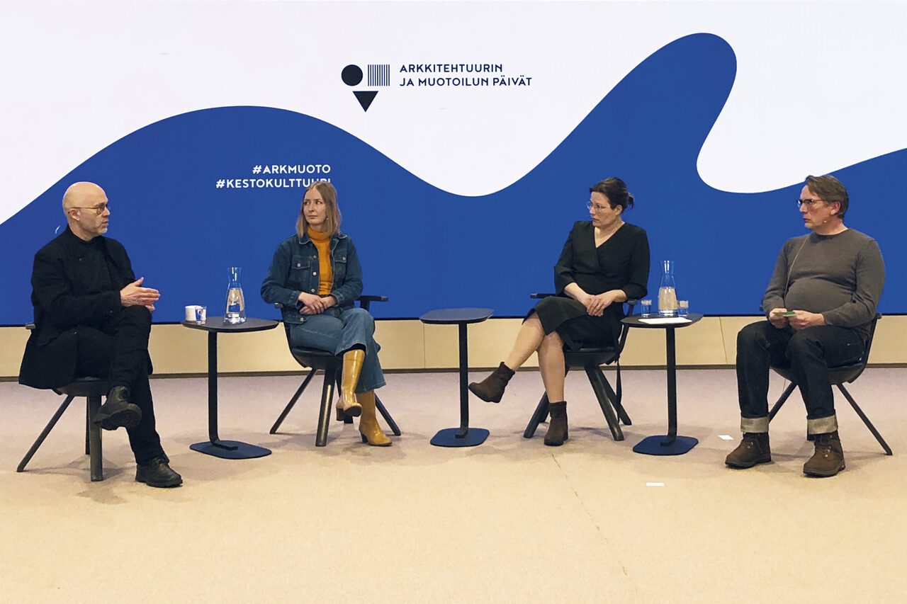 Neljä ihmistä istuu keskustelemassa, taustaseinällä suuri sininen aaltokuvio