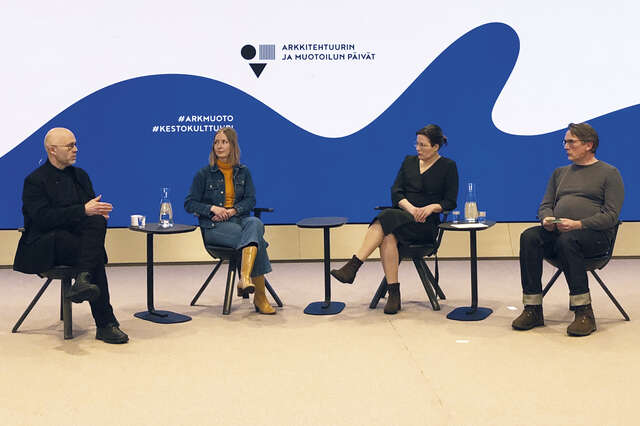 Neljä ihmistä istuu keskustelemassa, taustaseinällä suuri sininen aaltokuvio