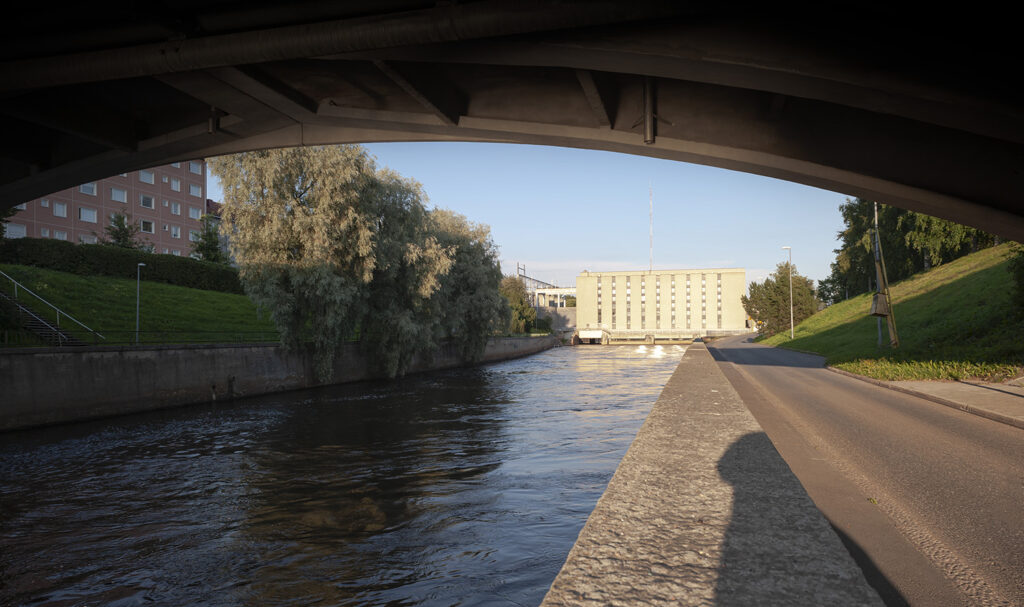 Näkymä kaupunkiympäristössä kaarevan siltarakenteen alta jokea pitkin kohti suurta vaaleaa rakennusta.